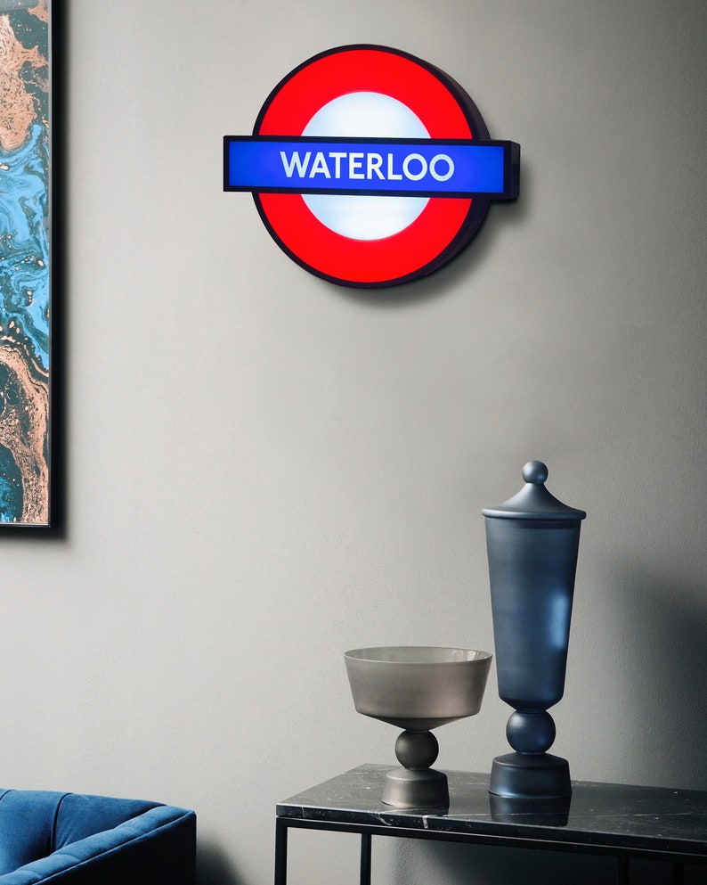 Peut inclure: Un panneau de m&eacute;tro londonien rouge et bleu avec le mot "WATERLOO" dessus. Le panneau est mont&eacute; sur un mur blanc.