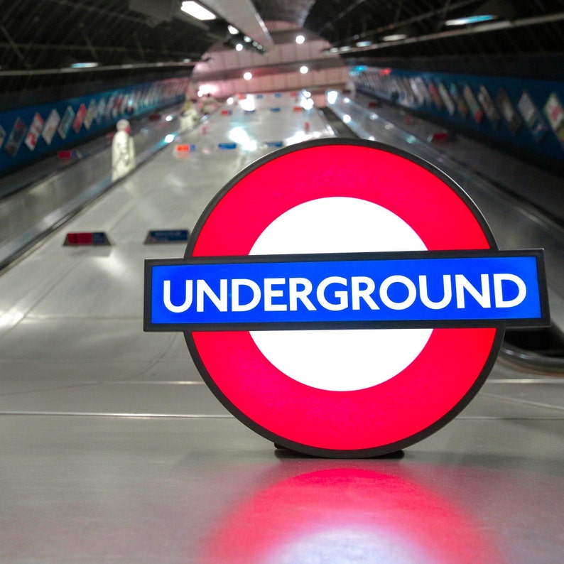 Peut inclure: Un panneau rond rouge et blanc avec le mot "UNDERGROUND" en lettres bleues sur une bande bleue. Le panneau est sur une surface grise.