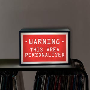 May include: A black-framed light box with a red illuminated panel. The panel displays the white text: "- WARNING - THIS AREA PERSONALISED". The light box is on a shelf above a collection of vinyl records.