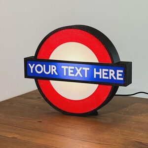 May include: A circular illuminated sign featuring a red ring and a central white circle. A horizontal blue rectangular bar crosses the center, displaying the white text "YOUR TEXT HERE". The sign has a black frame and a black power cord attached.