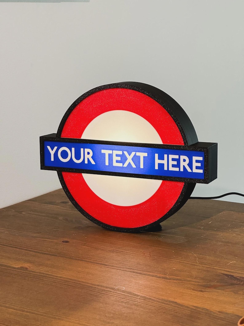 May include: A circular illuminated sign featuring a red ring and a central white circle. A horizontal blue rectangular bar crosses the center, displaying the white text "YOUR TEXT HERE". The sign has a black frame and a black power cord attached.