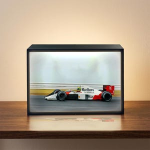 May include: A black framed display featuring a race car on a track. The car is white, red, and black, with "Marlboro" and "HONDA" branding. The display sits on a wooden surface.
