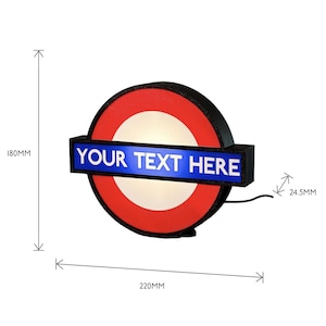 May include: A circular wall sign featuring a red ring and a central white circle, bisected by a horizontal blue rectangular bar with the white text "YOUR TEXT HERE". The sign measures 180mm in height, 220mm in width, and 24.5mm in depth.