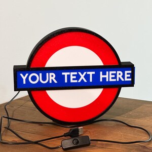 May include: A circular light-up sign featuring a red outer ring, a white center, and a horizontal blue bar across the middle. The blue bar displays the white text "YOUR TEXT HERE". The sign includes a black power cord with a USB connector and a switch.