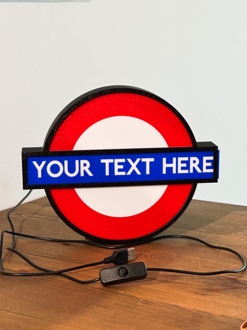 May include: A circular light-up sign featuring a red outer ring, a white center, and a horizontal blue bar across the middle. The blue bar displays the white text "YOUR TEXT HERE". The sign includes a black power cord with a USB connector and a switch.