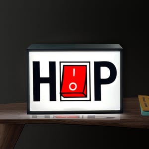 Op de afbeelding: Rechthoekige lichtbak met zwart frame met het woord "HOP" in vette zwarte letters. De "O" is vervangen door een rode schakelaar met een witte cirkel en een streepje. Op een houten oppervlak.
