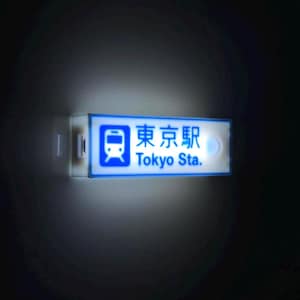 Puede incluir: Un letrero rectangular iluminado con el texto "東京駅 Tokyo Sta." en azul y blanco. El letrero presenta un icono de tren y probablemente es un indicador de estación. El fondo es oscuro.