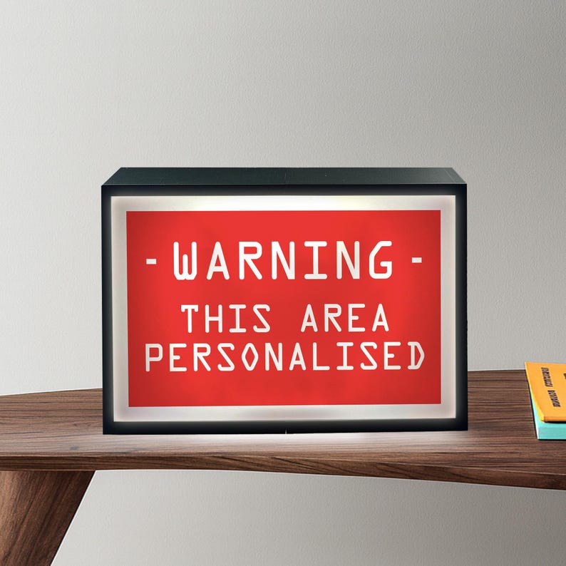 May include: A black rectangular light box with a red background and white text that reads "- WARNING - THIS AREA PERSONALISED". The light box is on a wooden surface.
