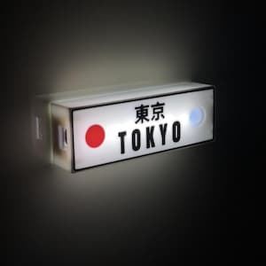 Può includere: Un'insegna rettangolare illuminata con la parola "TOKYO" in grassetto nero e caratteri giapponesi. Un cerchio rosso e un cerchio blu sono presenti sull'insegna. L'insegna è montata su una base bianca e si illumina intensamente.