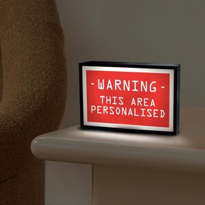 May include: A black rectangular light box with a red illuminated panel. The panel displays the white text: "- WARNING - THIS AREA PERSONALISED". The light box is on a white surface.