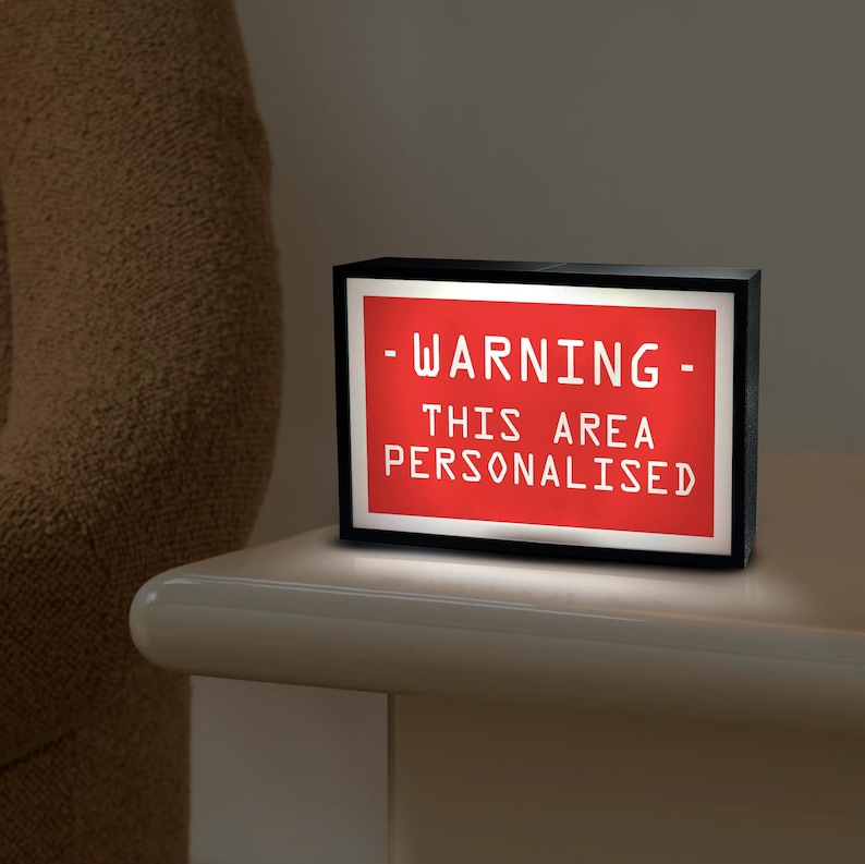 May include: A black rectangular light box with a red illuminated panel. The panel displays the white text: "- WARNING - THIS AREA PERSONALISED". The light box is on a white surface.