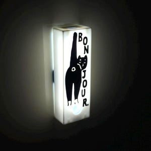 May include: A rectangular, illuminated wall light featuring a black cat illustration and the word "Bonjour" in black text. The light is white and mounted on a dark wall. The cat is depicted with its tail raised.