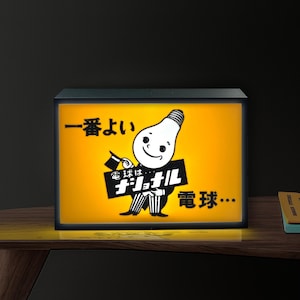 Op de afbeelding: Een verlicht bord in vintage-stijl met een zwart frame en een heldergele achtergrond. Het bord toont een stripfiguur van een gloeilamp die een bord met Japanse tekst vasthoudt. De tekst op het bord luidt "National" en "Light Bulb".