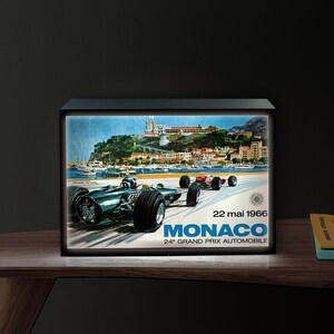 May include: Illuminated vintage Monaco Grand Prix poster. The poster features a green race car in the foreground, with other cars racing in the background. The poster has the text "Monaco 24° Grand Prix Automobile" and the date "22 mai 1966".