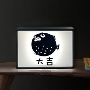 May include: A black rectangular light box with a white illuminated panel. The panel features a black cartoon pufferfish illustration and Japanese text. The light box is on a wooden surface.
