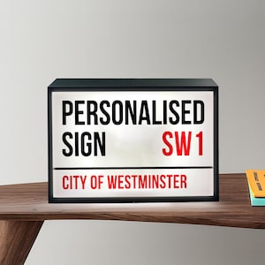 Op de afbeelding: Een zwarte rechthoekige lichtbak met de tekst "PERSONALISED SIGN SW1 CITY OF WESTMINSTER" in zwart en rood. De lichtbak staat op een houten oppervlak.