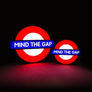 Peut inclure: Deux panneaux du m&eacute;tro londonien rouge et blanc avec du texte bleu qui dit "MIND THE GAP".
