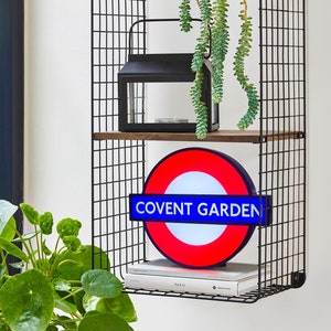 Peut inclure: Un panneau de m&eacute;tro londonien rouge, blanc et bleu avec les mots "Covent Garden" dessus. Le panneau est suspendu &agrave; une &eacute;tag&egrave;re en fil de fer noir avec une lanterne noire et une plante verte suspendue au-dessus.