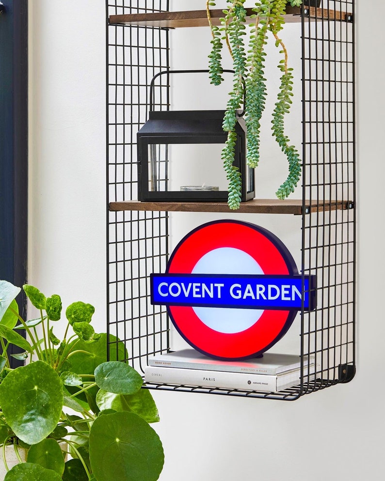 Peut inclure: Un panneau de m&eacute;tro londonien rouge, blanc et bleu avec les mots "Covent Garden" dessus. Le panneau est suspendu &agrave; une &eacute;tag&egrave;re en fil de fer noir avec une lanterne noire et une plante verte suspendue au-dessus.