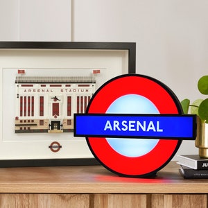 Peut inclure: Une enseigne lumineuse rouge et bleue avec le mot "ARSENAL" en lettres blanches. L'enseigne est en forme de panneau du m&eacute;tro londonien.