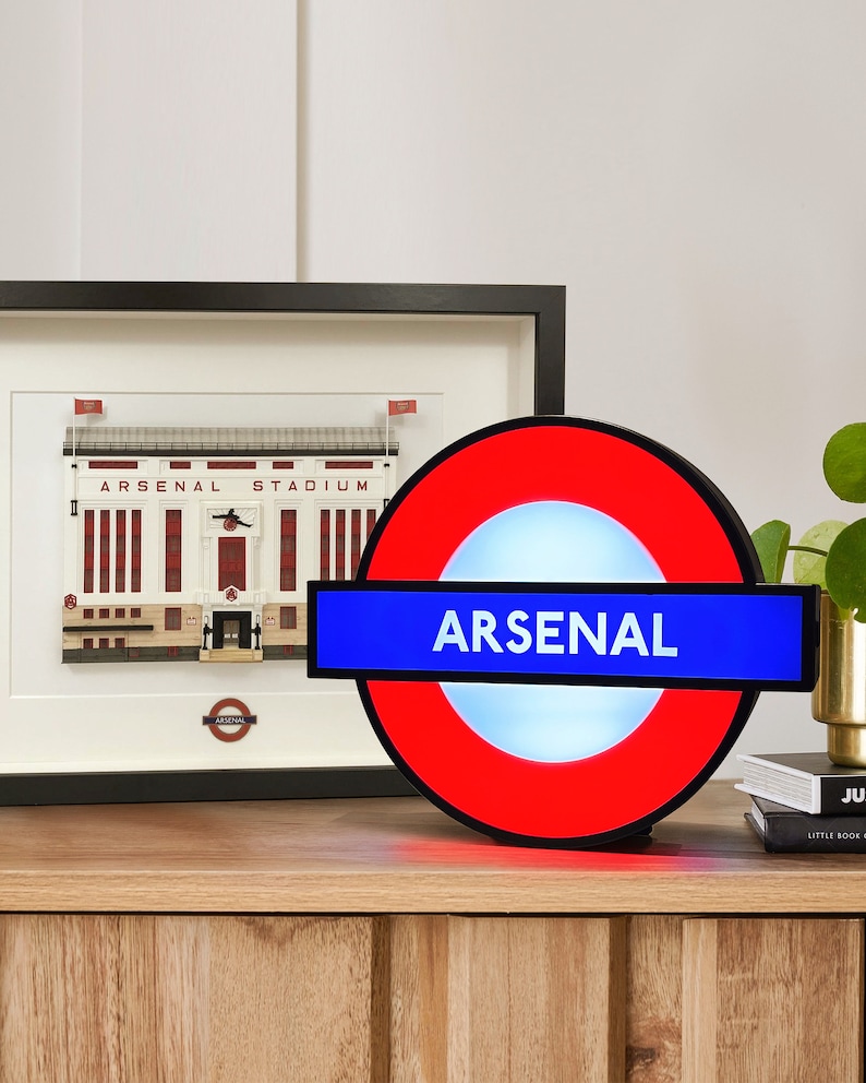 Peut inclure: Une enseigne lumineuse rouge et bleue avec le mot "ARSENAL" en lettres blanches. L'enseigne est en forme de panneau du m&eacute;tro londonien.