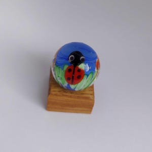 May include: A glass marble with a blue background and a painted ladybug design. The marble is on a small wooden stand.