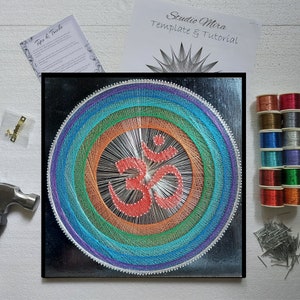 Op de afbeelding: Een regenboogkleurig string art ontwerp van het Om-symbool op een zwarte achtergrond. Het ontwerp is ingelijst en bevat een set kleurrijke borduurgaren, spijkers en een hamer.