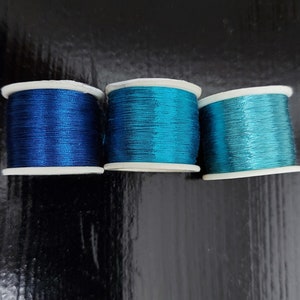 May include: Three spools of metallic thread in shades of blue. The spools are arranged in a row from left to right, with the darkest blue spool on the left and the lightest blue spool on the right.