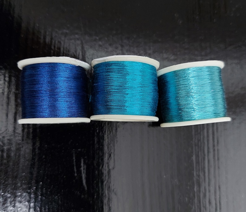 May include: Three spools of metallic thread in shades of blue. The spools are arranged in a row from left to right, with the darkest blue spool on the left and the lightest blue spool on the right.