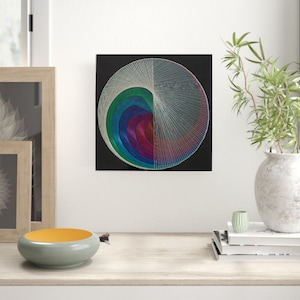 May include: A colourful string art piece with a circular design on a black background. The design features a spiral of colourful threads, with a white thread creating a radial pattern.