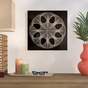 Pode incluir: Uma tela quadrada preta com um design geométrico de arte de corda branca. O design é um círculo com uma série de ovais e linhas sobrepostos.