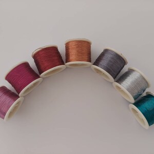 May include: Seven spools of thread in various colours, including purple, pink, red, copper, silver, and teal, arranged in a semi-circle on a white surface.