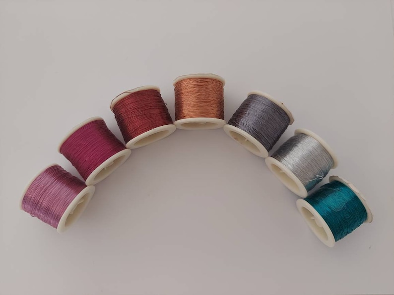 May include: Seven spools of thread in various colours, including purple, pink, red, copper, silver, and teal, arranged in a semi-circle on a white surface.