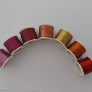 May include: A rainbow of colours in spools of thread, arranged in a semi-circle on a white background. The colours are pink, purple, red, orange, yellow, and gold.