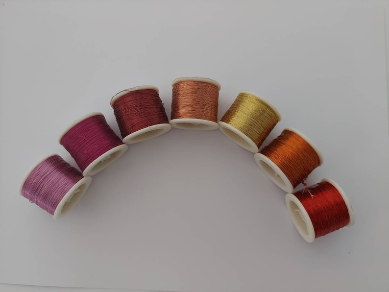 May include: A rainbow of colours in spools of thread, arranged in a semi-circle on a white background. The colours are pink, purple, red, orange, yellow, and gold.