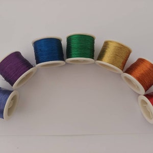 May include: Seven spools of thread in a rainbow of colours, arranged in a semi-circle on a white surface. The colours are blue, green, gold, orange, red, purple, and a lighter blue.