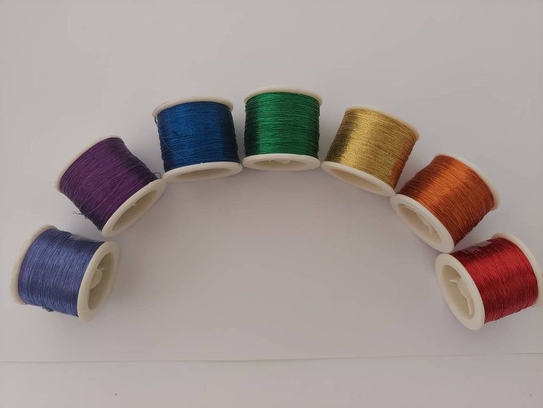 May include: Seven spools of thread in a rainbow of colours, arranged in a semi-circle on a white surface. The colours are blue, green, gold, orange, red, purple, and a lighter blue.