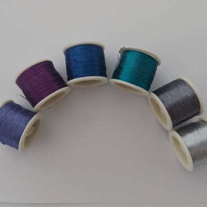 May include: Six spools of thread in various colours, including purple, blue, teal, silver, and grey. The spools are arranged in a semi-circle on a white surface.