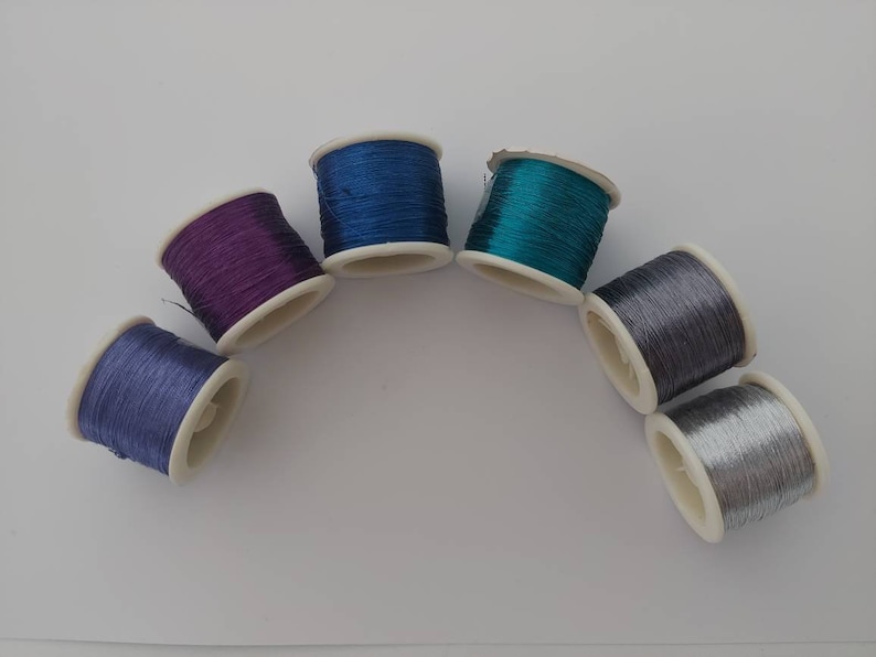 May include: Six spools of thread in various colours, including purple, blue, teal, silver, and grey. The spools are arranged in a semi-circle on a white surface.