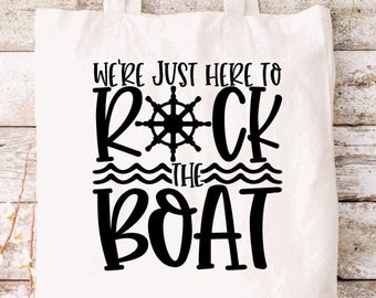 May include: A white canvas tote bag featuring black text that reads WE'RE JUST HERE TO ROCK THE BOAT. The letter O in ROCK is replaced by a black ship's wheel graphic, and two wavy lines are positioned between the words ROCK and THE.