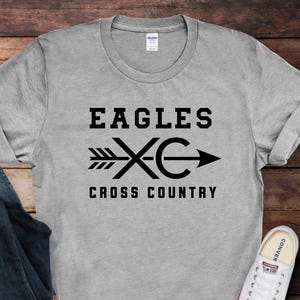May include: A heather gray short-sleeve t-shirt featuring a black graphic print that reads EAGLES XC CROSS COUNTRY with an arrow design. The shirt is displayed alongside dark blue denim jeans and a pair of white canvas sneakers.