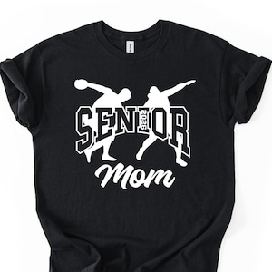 May include: Black t-shirt with a white graphic. The graphic features two stylized figures in motion, the word "SENIOR" with the year "2026" and the word "Mom" in a script font. The shirt has short sleeves and a crew neck.