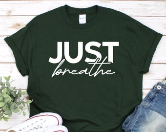 May include: A dark forest green short-sleeved t-shirt featuring the white text "JUST" in a bold sans-serif font above the word "breathe" written in a white cursive script. The shirt has a crew neckline and a Gildan brand label at the collar.