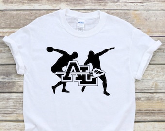 May include: A white short-sleeve t-shirt featuring a black graphic of two athletes in motion. One athlete prepares to throw a discus, while the other prepares to throw a shot put. The letters AL are centered between them with a spearhead icon.