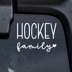 May include: A white vinyl decal sticker featuring the word HOCKEY in a tall, sans-serif font stacked above the word family in a cursive script font, with a small heart icon at the end of the word family, applied to a dark surface.