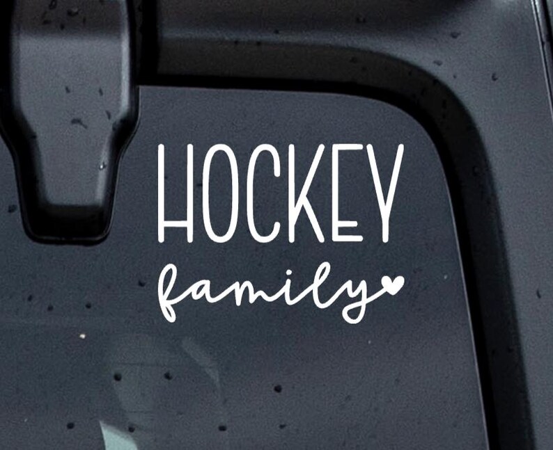May include: A white vinyl decal sticker featuring the word HOCKEY in a tall, sans-serif font stacked above the word family in a cursive script font, with a small heart icon at the end of the word family, applied to a dark surface.