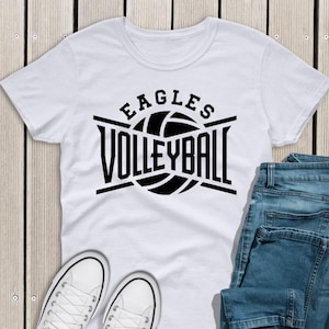 May include: White t-shirt with the text "EAGLES VOLLEYBALL" in black, featuring a volleyball graphic. The shirt is paired with blue jeans and white sneakers, set against a wooden background.