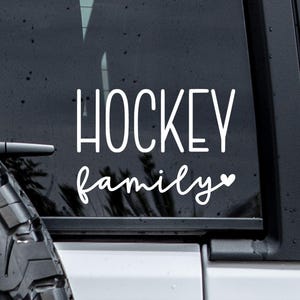 May include: A white vinyl decal applied to a dark vehicle window featuring the word HOCKEY in a tall, sans-serif font above the word family in a cursive script font, finished with a small heart icon at the end of the text.