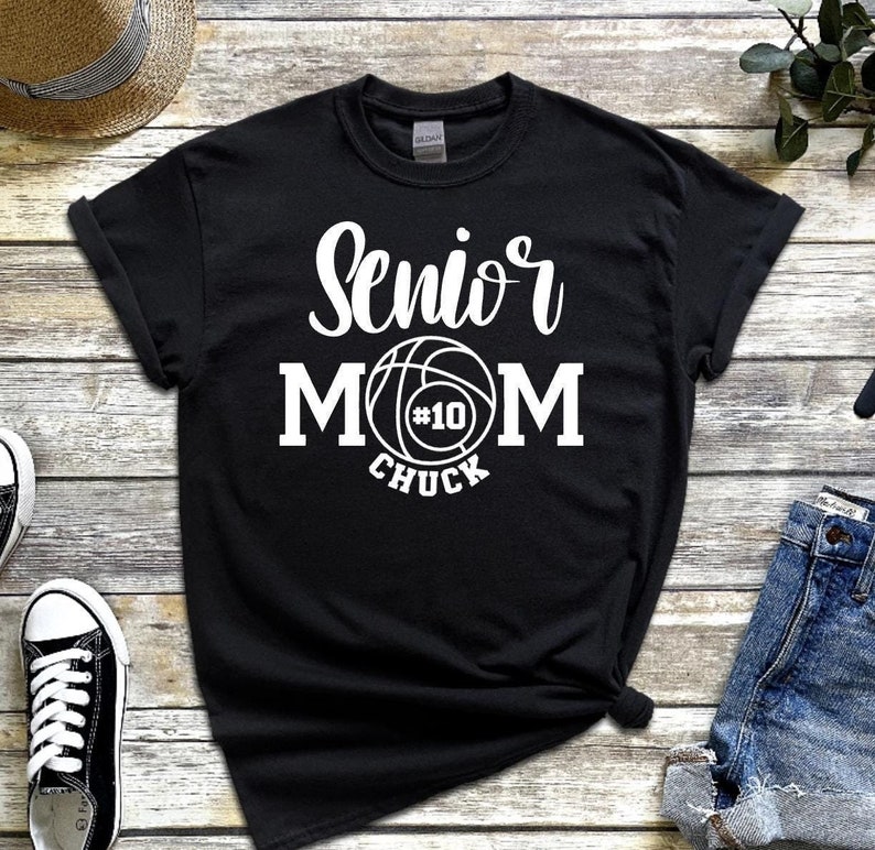 May include: Black t-shirt with white text that reads "Senior Mom" with a basketball graphic and the number "#10" and the word "Chuck" underneath.