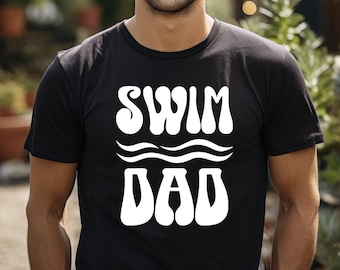 May include: A black short-sleeved t-shirt featuring the white text SWIM DAD in a bold, wavy font. Between the two words are two horizontal white wavy lines representing water. The shirt has a crew neckline and a standard fit.